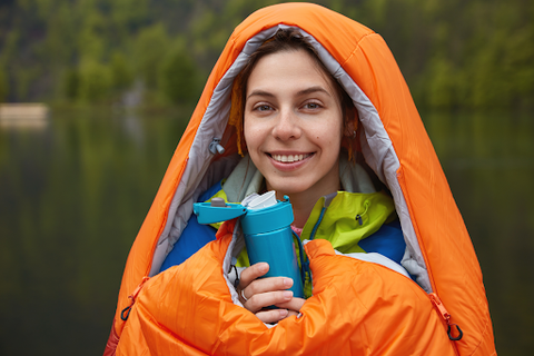 A lady standing in her orange bivy drinking out of a blue flask with a lake and trees in the background