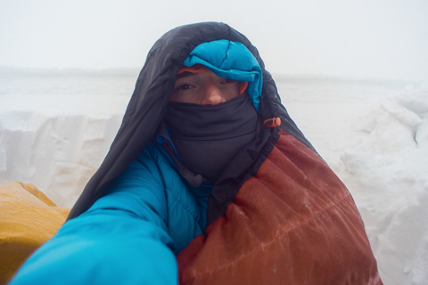 man outdoors in snow with sleeping bag over head
