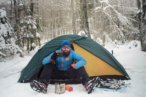 man outside tent in snow in warm clothes and warm drink