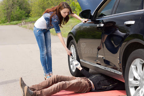 A lady standing by her car handing the man a tool as he is lying underneath the car