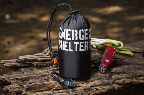 An Emergency USA emergency shelter in its bag on a log with a army knife and compass next to it