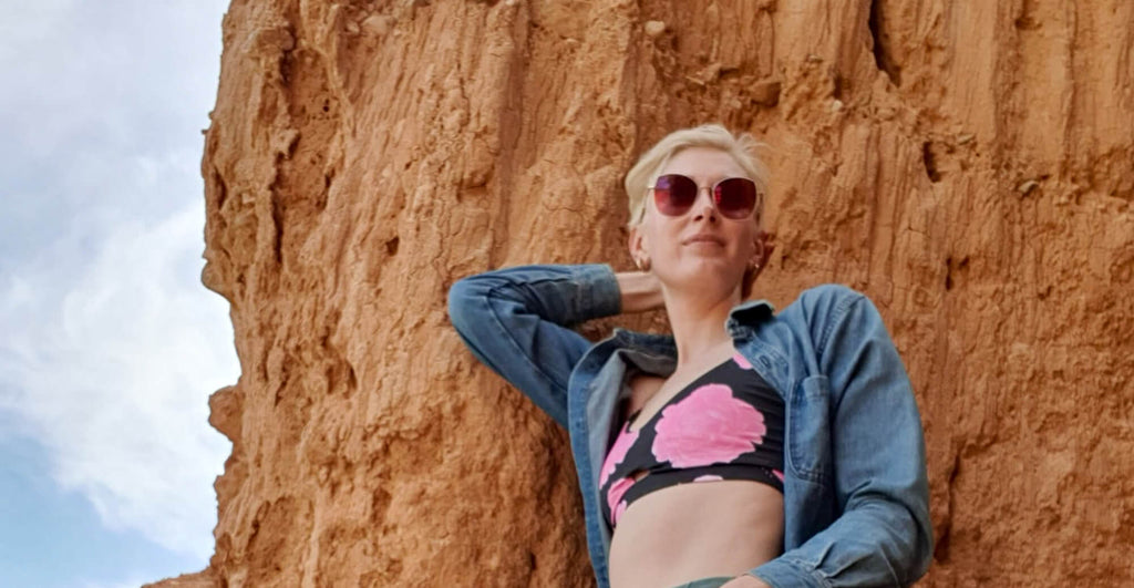 Woman in Ibiza leaning against a rock wall at the beach, wearing a pink and green sports bra with a floral motif and a denim shirt.