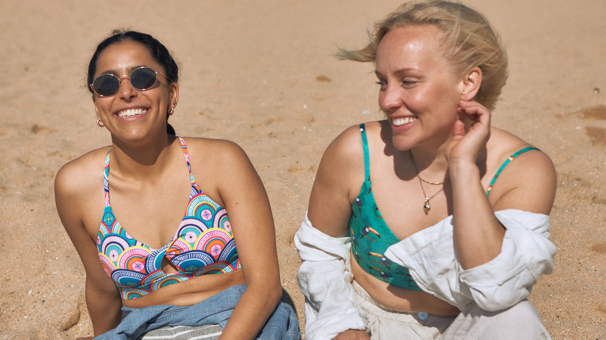 two women sitting next to each other on the beach and laughing. One is wearing a multicolor sports bra with a colorful, geometric design. The other is wearing a green sports bra with a hand-painted Toucan print and white and cream linen shirt and pants.