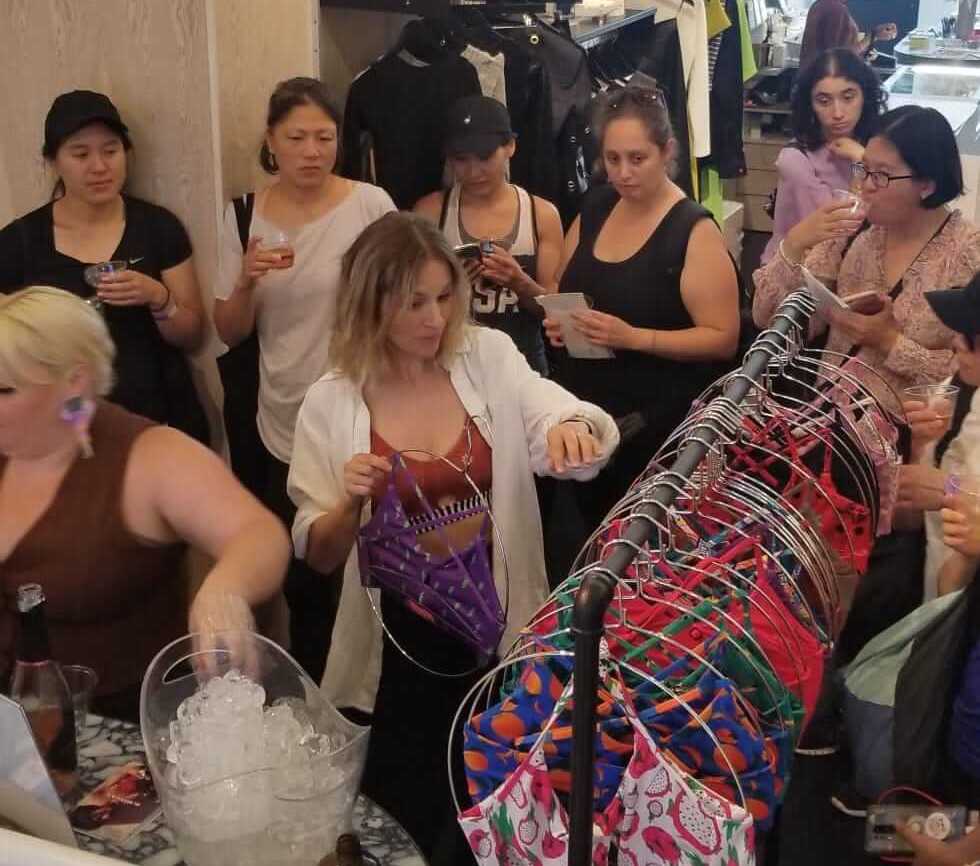 Women at a fashion sports bra brand launch in NYC, designer is showing various bra tops on hangers to a crowd.