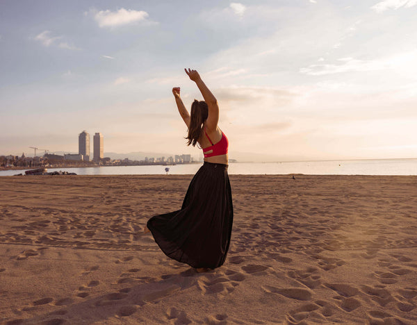 Woman dancing alone at sunset on the beach wearing a black flowy dress and pink sports bra
