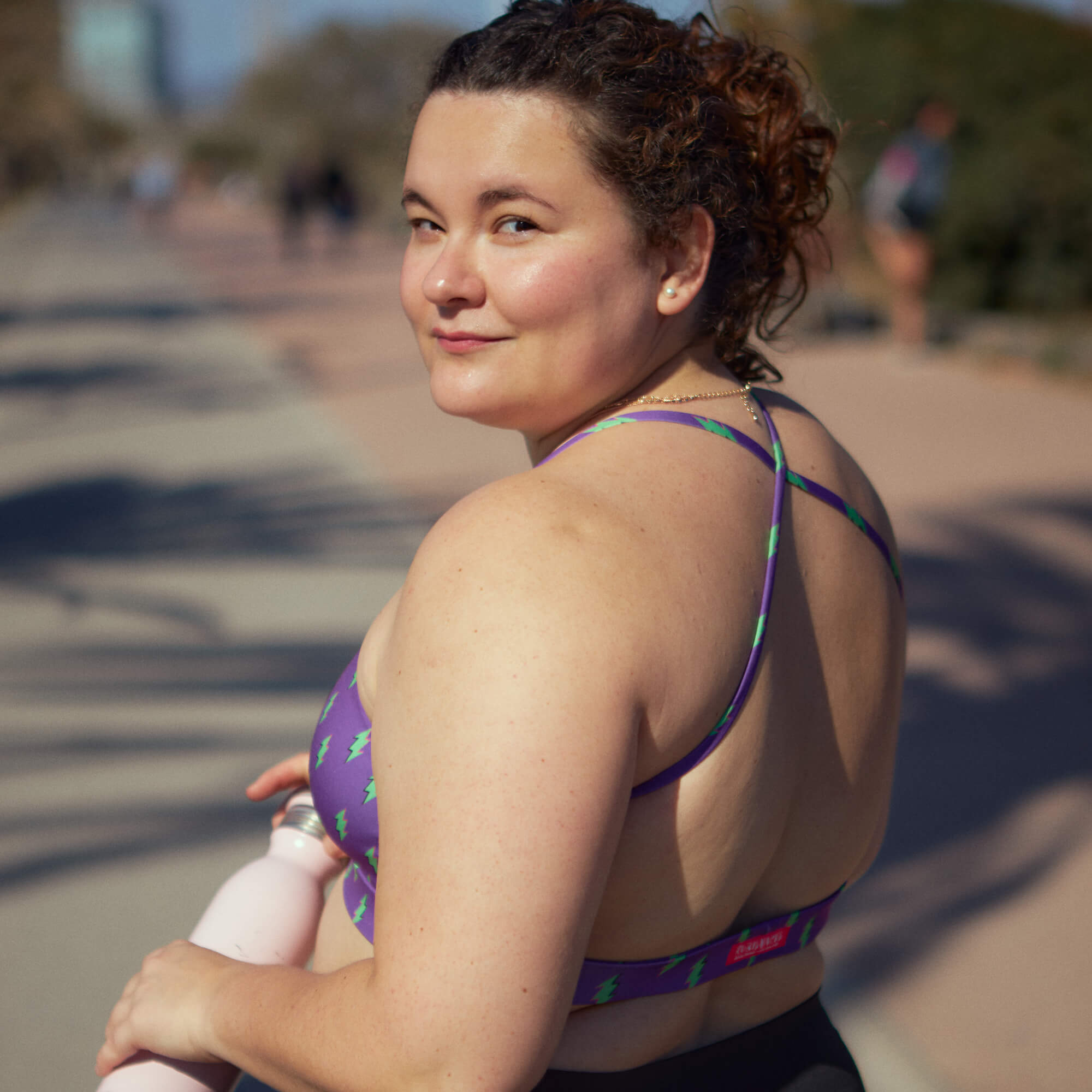 Woman looking back over her shoulder, wearing a lightning bolt sports bra in purple and holding a water bottle.