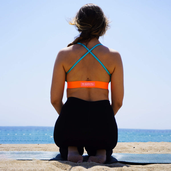 A woman with her back to us in a yoga pose, looking into the distance at a beach. She is seating on her legs and wears a racerback sports bra in light blue and bright neon orange. Her hair is in a loose braid.