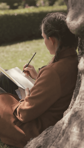 woman sitting against a tree with an open book, sketching