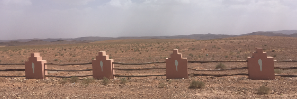 Road side picture of a fence in Morocco with terracotta earth posts in a geometric shape and wooden beams in between. The landscape is desert and mountains.