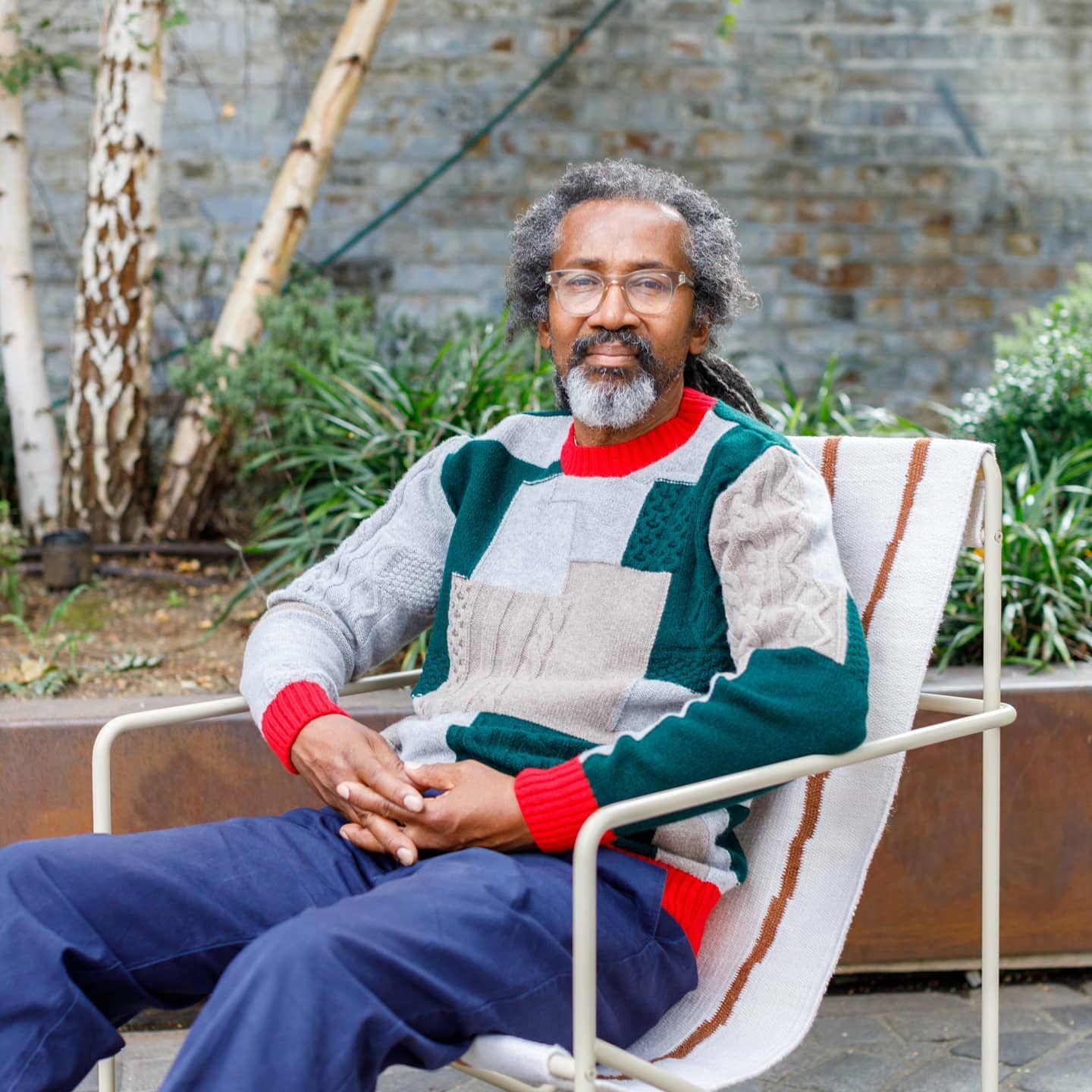 Herbie sits in a garden, wearing a colorful patchwork sweater. 