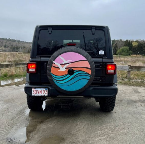 Seagulls flying in a beach scene with waves and the ocean on a spare tire cover for black Jeep Wrangler. Spare tire cover is in shades of purple and blue