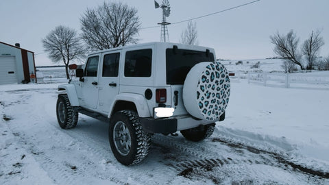 White Jeep Wrangler parked in the snow with a white vinyl spare tire cover that has a leopard design.