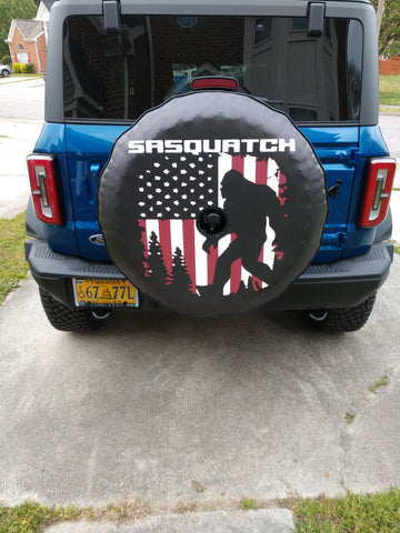 Sasquatch or big foot silhouette walking in front of an American flag with the word Sasquatch on top, spare tire cover design on a blue Ford Bronco