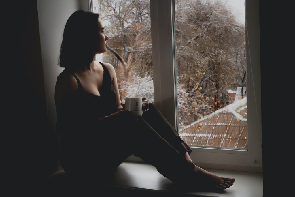 Femme assise sur le rebord de la fenêtre qui regarde dehors avec une tasse de café à la main