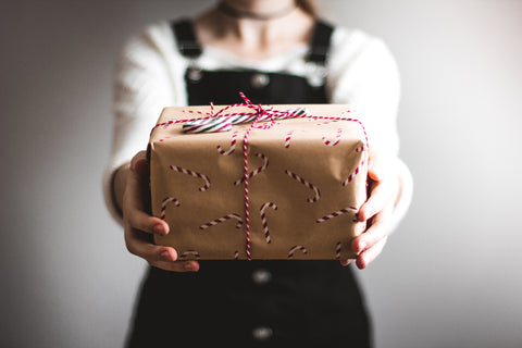 Young woman in black overalls extending a gift wrapped in brown kraft paper and tied with red-and-white yarn