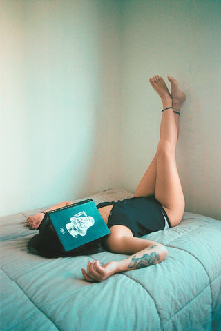 Young woman in underwear and tank top on bed with legs propped against wall and book covering face