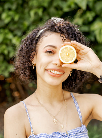 Young woman with dark curly hair and blue sundress holding cut lemon in front of eye