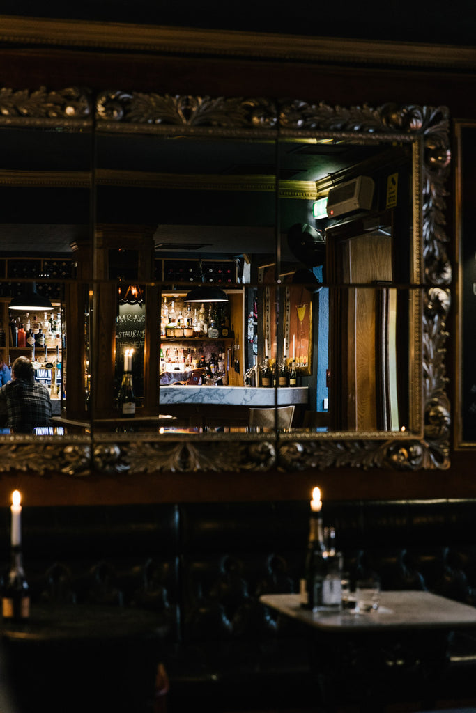 Image of an ornamental golden mirror, reflecting a bar with atmospheric lighting.