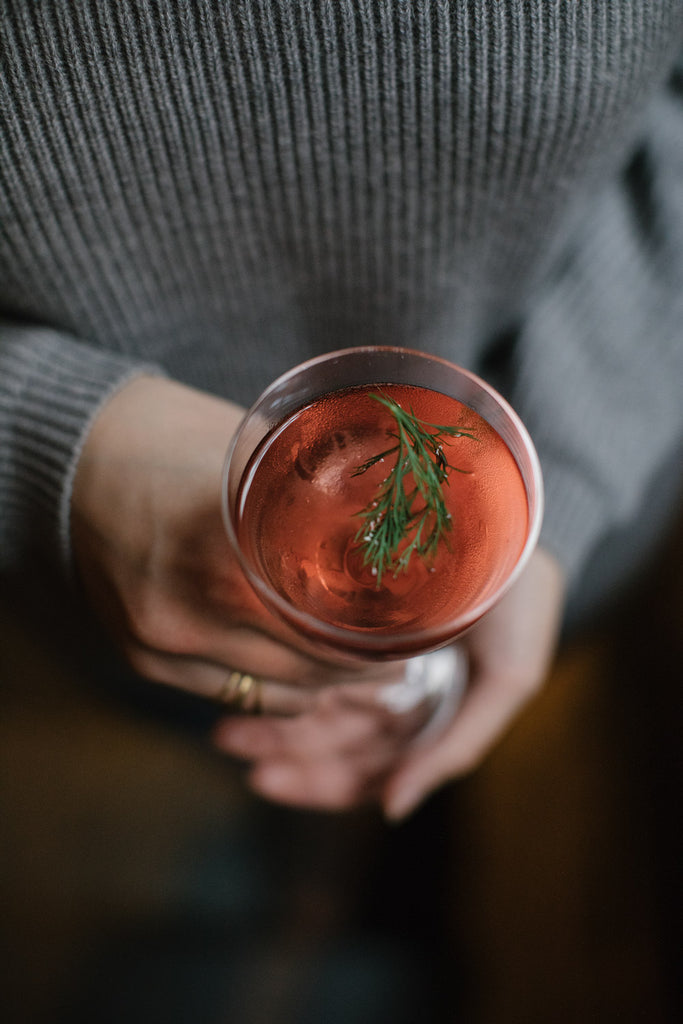 A close-up image of a red cocktail drink with an herb on top, held by a person in a grey sweater.