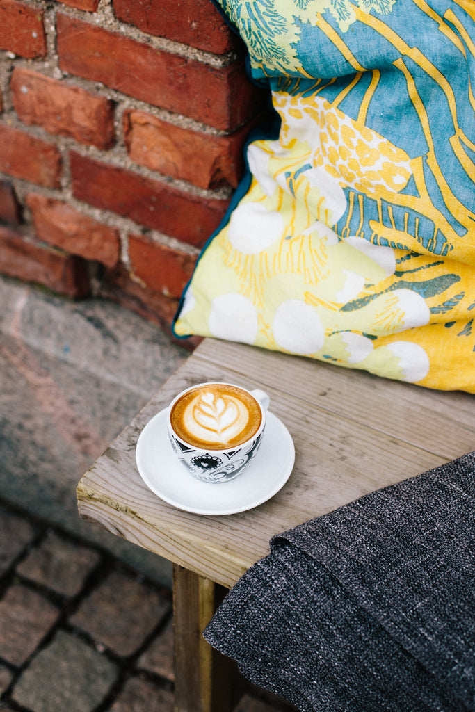 A photograph of a coffee cup with latte art, on a bench next to a red tile wall and a colorful cushion.