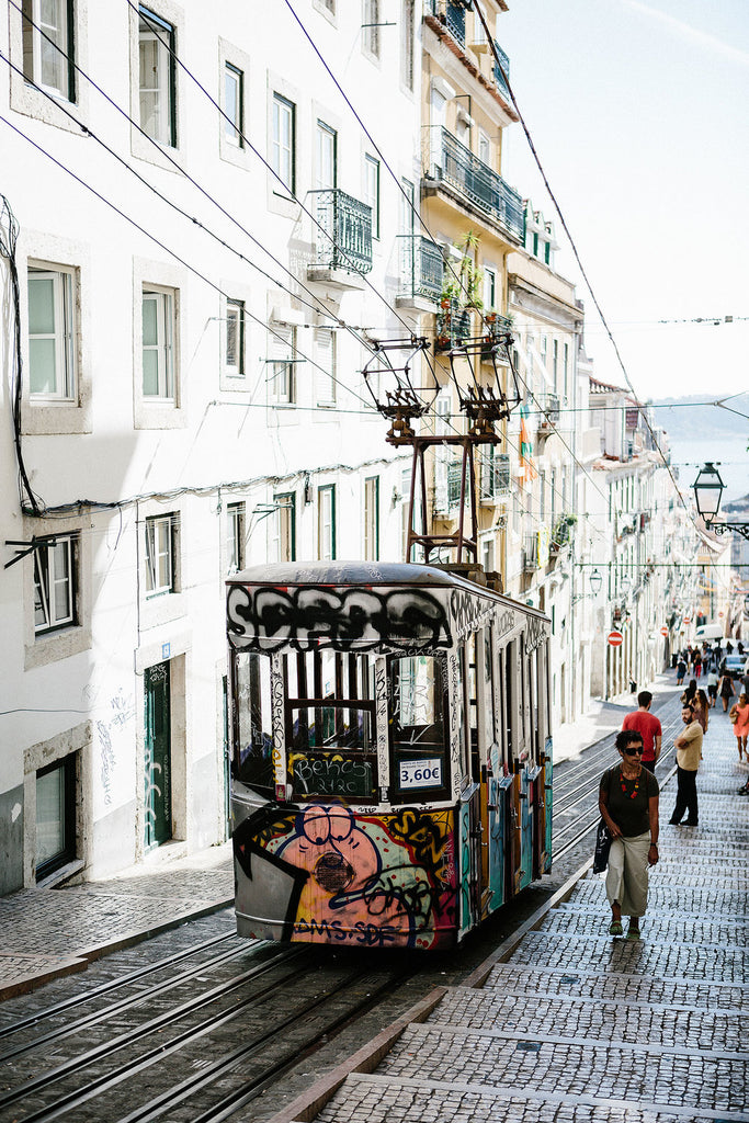 Image of the Lisbon street view: a tram decorated with graffiti moving in front of white buildings and a distant view to water.