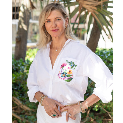 Image of the founder Sandra Greenberger standing in her shop front wearing a white top with flowers embroidered on the right hand side.