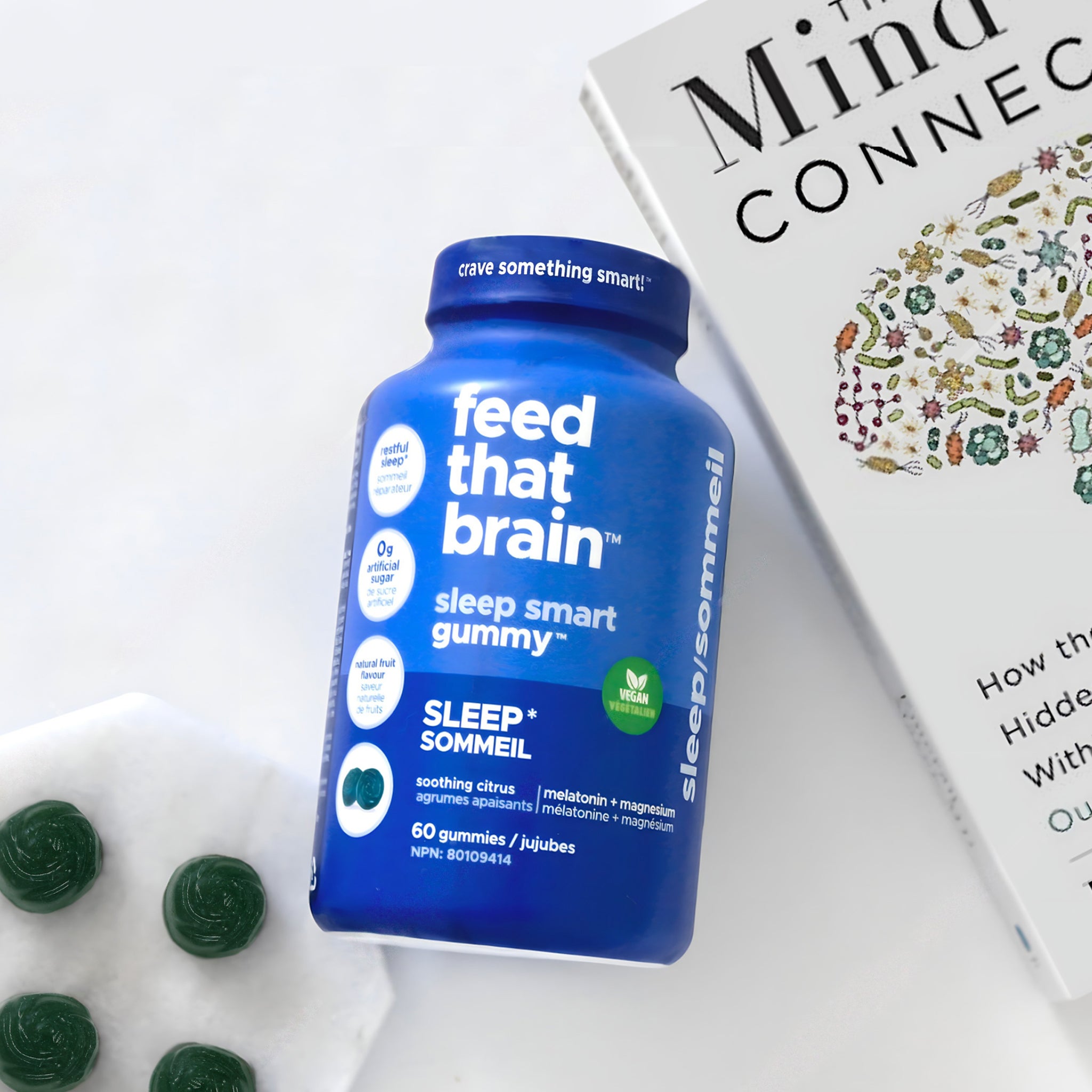 blue feed that brain sleep gummy bottle laying on a table, beside a mind connection book and a white plate with loose blue sleep gummies sprawled on it.