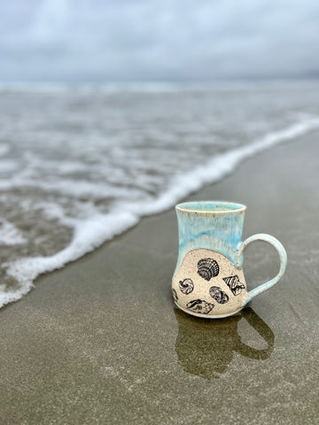 Handmade mug with seashells and sea foam on beach by waves