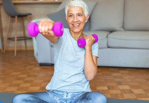 middle aged woman working out in her living room. She is wearing a blue tshirt and yoga pants and holding two pink dumbbells. 