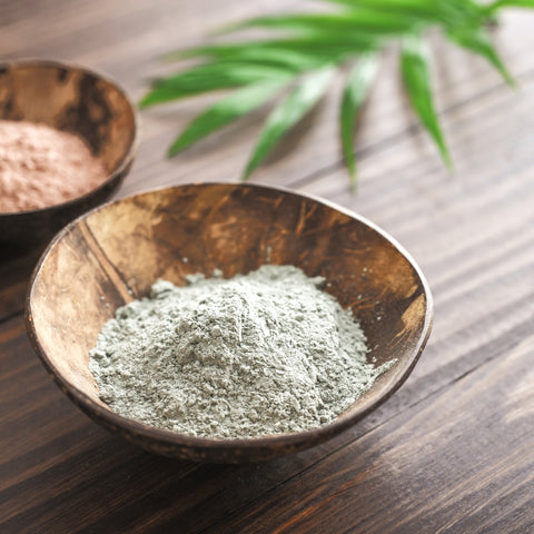 Two wooden bowls of French Green and French Pink Clays on a natural wood surface with a fresh green leaf beside them