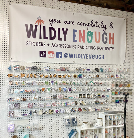 White pegboard wall with rows of clear shelves with stickers, under a vinyl hanging sign that says Wildly Enough.