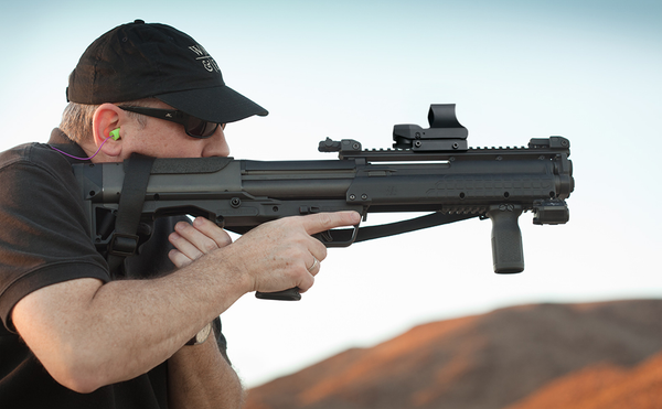 A marksman using a rifle with an attached reflex sight for improved aiming stability in a field setting.