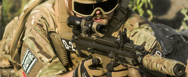 Military operative in field gear aiming with a scoped tactical rifle, highlighting the use of optical sights in combat.