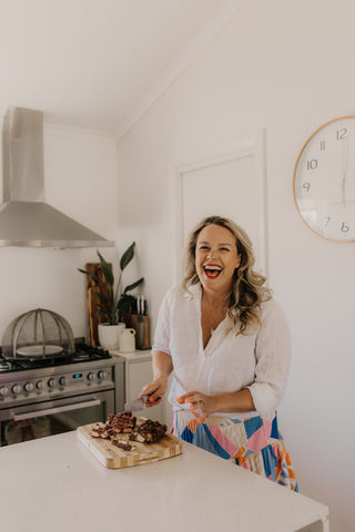 tash the owner is in a kitchen in mudgee nsw cutting up rocky road. she is laughing at the camera, wearing a white shirt and a multicoloured skirt.