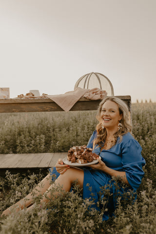 Tash is sitting in a field of lucerne crop, in front of a huge old table, in a blue dress, with a huge smile, eating a huge platter of her own Pebbly Path rocky road