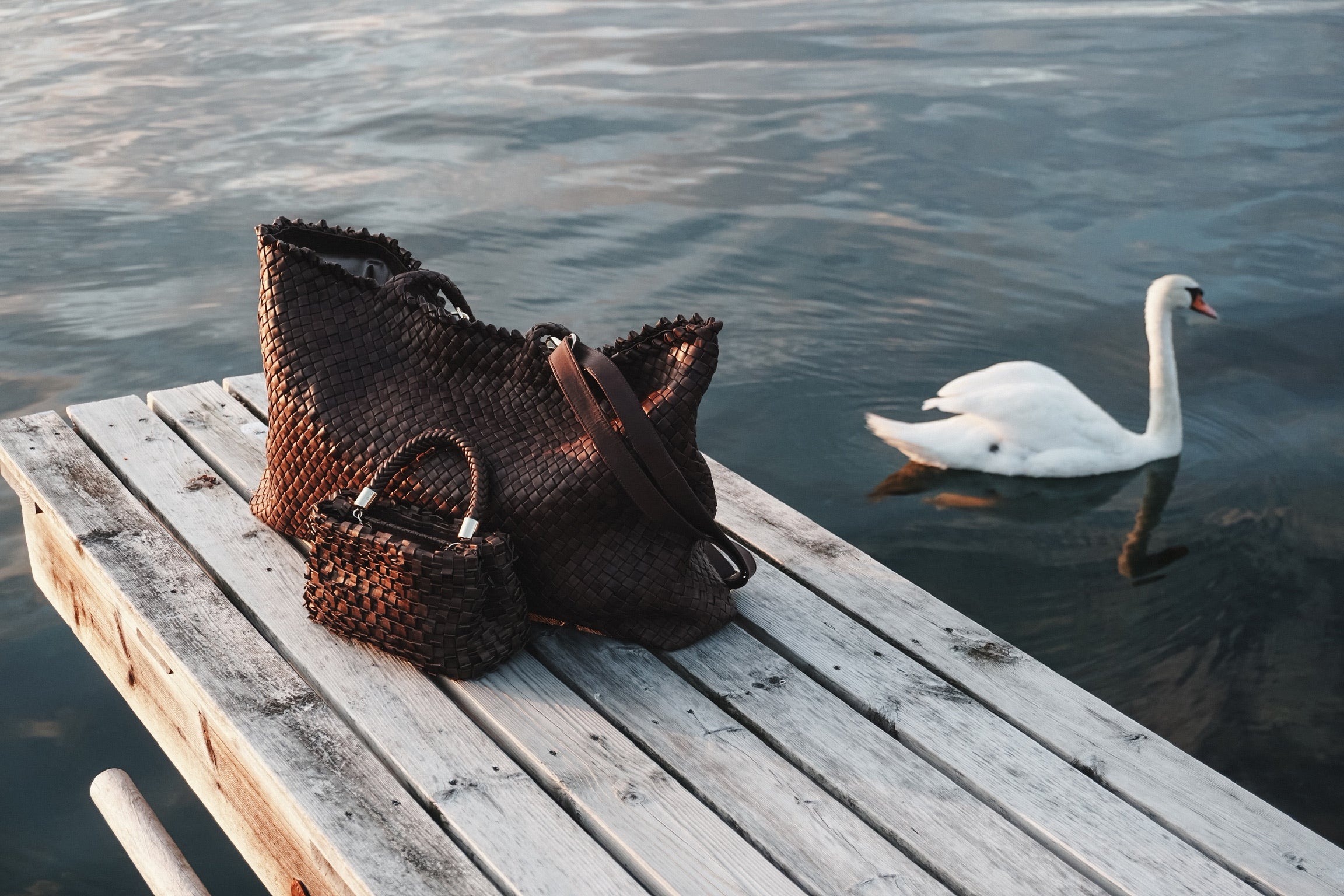Ribichini håndvesker i flettet skinn på bryggekant ved vannet, med en svane i bakgrunnen ved solnedgang.