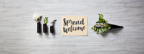 Three little vases with flowers lying next to a wooden card that reads "Spread Welcome" followed by another vase lying perpendicular with greenery