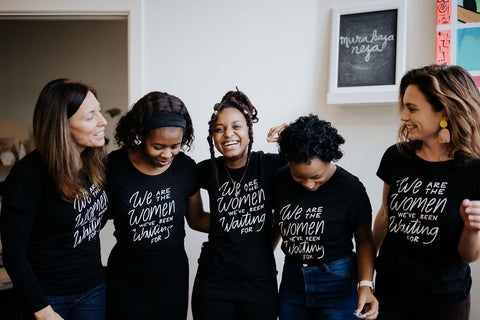 Five women of different ages and races standing together and laughing. All are wearing black shirts with white text that reads "We are the Women We've Been Waiting For"