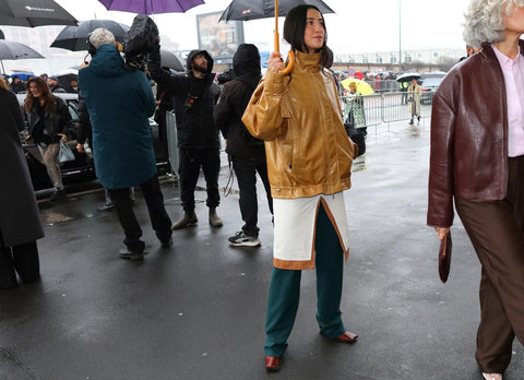 woman holding an umbrella, wearing a beige leather jacket and a white dress over green pants