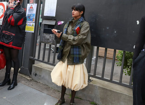 woman with a long dark olive jacket, long tartan blue scarf and a balloon ivory skirt