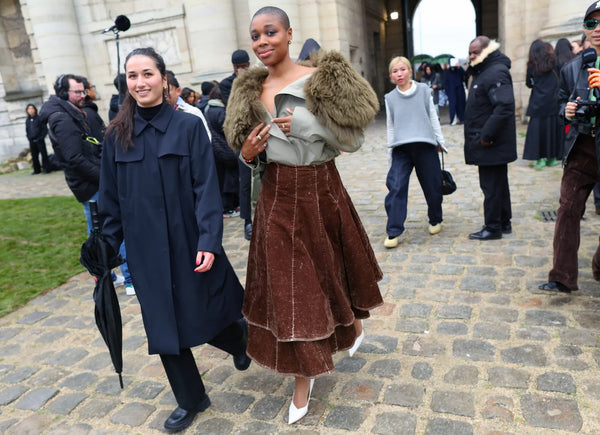 woman with black suit accompanied by woman with a mint shirt with big faux fur collar, brown skirt and white pumps