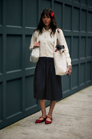 woman with cream cardigan, full skirt, red ballet flats and a small dog in a shopping bag