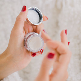 Mains de femme aux ongles avec du vernis rouge. La main gauche tient un pot de crème visage, et la main droite prélève dans ce pot.