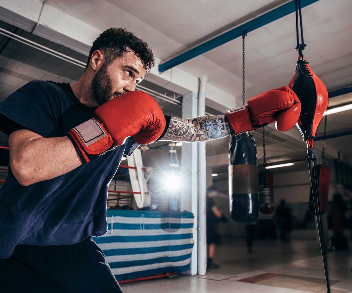 Light-skinned, bearded man with 1 tattoo sleeve wearing boxing gloves and punching a speed bag.