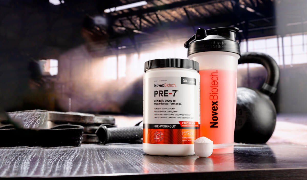 A tub of PRE-7 preworkout powder, a scoop of the powder, and a shaker bottle mixed with the powder in the foreground; gym equipment in the background.