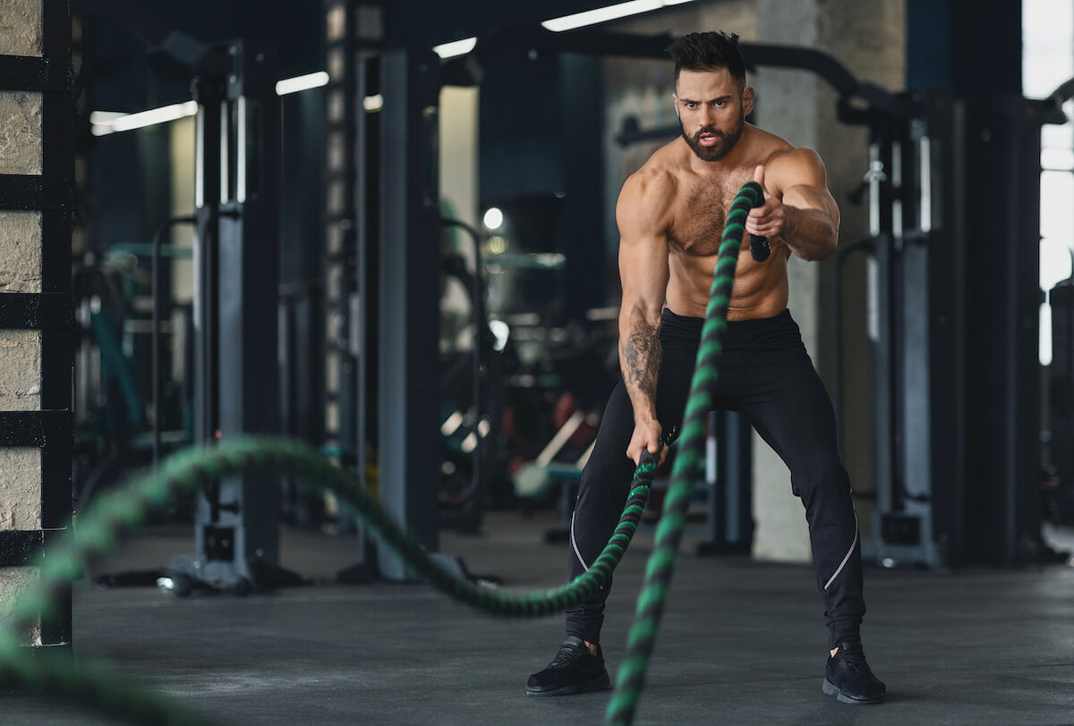 An athletic, light-skinned man wearing workout pants with his shirt off as he concentrates on a ropes workout.