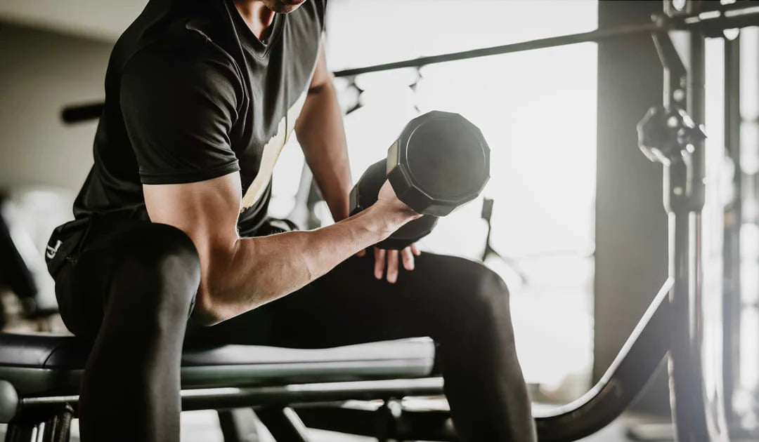 Man sitting on a weight bench curling a barbell with one arm