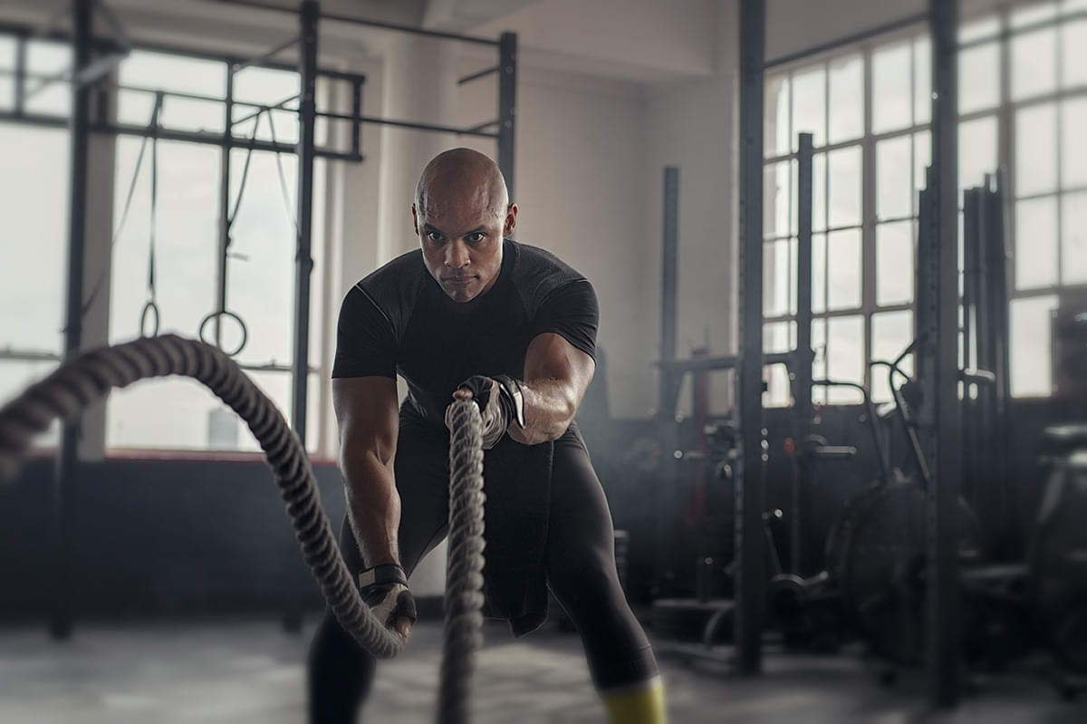 Dark-skinned man using battle ropes in a gym.