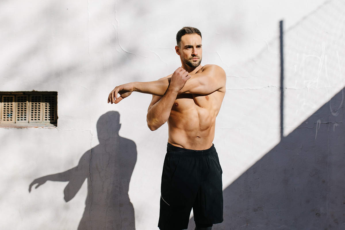 White man in workout shorts standing in front of a white wall stretching before a workout