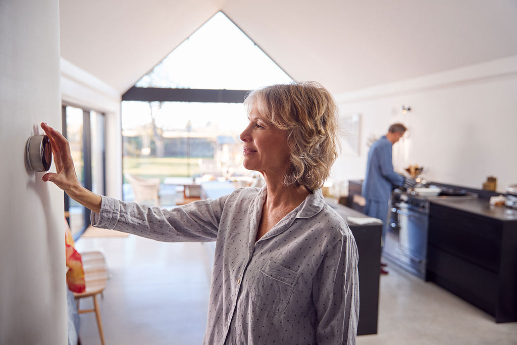 older woman turning up thermostat while husband cooks breakfast